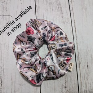May include: Pink satin scrunchie with a print of musical instruments, typewriters, and other objects. The scrunchie has a small tag that says "T.S. scrunchie available in shop".
