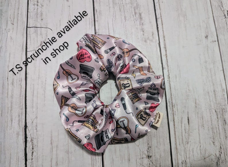 May include: Pink satin scrunchie with a print of musical instruments, typewriters, and other objects. The scrunchie has a small tag that says "T.S. scrunchie available in shop".