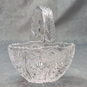 May include: A clear glass basket featuring a frosted rose motif on the sides and a textured leaf pattern on the arched handle. The body of the basket displays a faceted diamond-cut design with a scalloped rim, crafted from transparent glass.