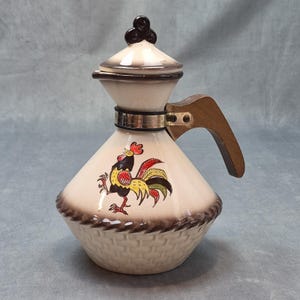 May include: A cream-colored ceramic pitcher with a conical body and a matching lid featuring a dark brown finial. The base has a molded basket-weave texture with brown shading. A colorful rooster illustration is centered on the front, and a wooden handle is attached with a metal band.