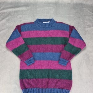 Könnte beinhalten: Ein langärmeliger Strickpullover mit horizontalen Streifen in den Farben Blau, Magenta und Dunkelgrün. Das Kleidungsstück verfügt über einen gerippten Rundhalsausschnitt, gerippte Bündchen und einen gerippten Saum mit durchgehendem Streifenmuster.