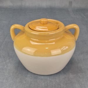 May include: A two-handled ceramic jar with a matching lid, featuring a two-tone design with a glossy mustard yellow glaze on the upper portion and a matte off-white finish on the lower body, complete with two curved side handles for easy gripping.