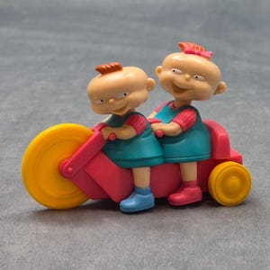 May include: A plastic toy figurine featuring two cartoon characters with peach skin tones, orange hair, and teal outfits riding a pink tricycle with a large yellow front wheel and a smaller yellow rear wheel. The characters have distinct facial expressions.