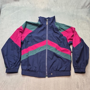 Peut inclure: Un coupe-vent bleu marine doté d'un col montant, d'une fermeture éclair intégrale sur le devant et de manches longues à poignets élastiques. La veste est rehaussée de bandes diagonales contrastées en magenta vif et vert forêt sur la poitrine.
