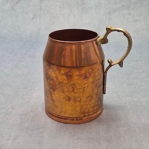 May include: A hammered copper mug with a cylindrical body and a curved, ornate brass handle. The exterior surface features a textured, dimpled finish with varying shades of metallic orange and brown, contrasting with the smooth, polished brass handle.