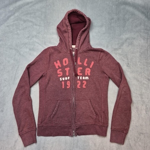 May include: A maroon zip-up hoodie featuring a hood with a dark interior lining. The front displays the text Hollister Surf Team 19 22 in raised, textured coral and white lettering. The garment includes long sleeves and two front pockets.