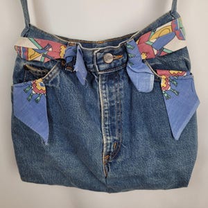 May include: A blue denim tote bag with a colorful patterned fabric strap and trim. The bag is made from repurposed denim and has a unique, handmade look.