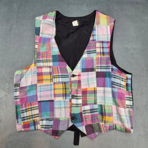 May include: A multi-colored patchwork vest with a black back and lining. The vest features a V-neck, button closure, and a variety of plaid patterns in shades of pink, purple, yellow, and blue. The vest is a unique piece of clothing.