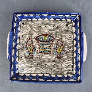 May include: A square ceramic serving tray with handles, featuring a white background with a black grid pattern. The center displays a colorful mosaic-style design of a basket and two fish. The border has a blue scalloped pattern on white.