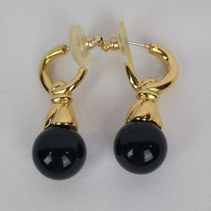 May include: A pair of gold-tone earrings with a post back. Each earring features a gold-tone hoop design, a gold-tone knot detail, and a large, glossy black sphere. The earrings are displayed against a white background.