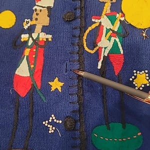 May include: Blue knit sweater with embroidered toy soldier figures in red, green, and white uniforms. The sweater features yellow stars and circles, and black trim along the button closure. The figures are standing on brown and green platforms.