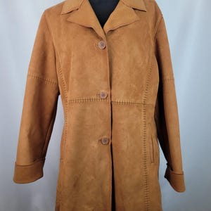 May include: A tan suede-textured coat featuring a collared neckline, long sleeves with cuffs, and a front button closure with three matching buttons. The garment includes decorative whipstitch detailing along the vertical seams and waistline.