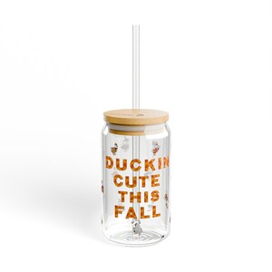Może przedstawiać: Przezroczysty szklany kubek z bambusową pokrywką i słomką. Na kubku widnieje napis "DUCKIN CUTE THIS FALL" pomarańczowymi literami. Małe ilustracje kaczek są rozrzucone po szkle.