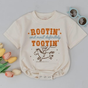 May include: Cream-colored baby onesie with the text "Rootin' and most definitely Tootin'" in orange and blue. The graphic features a cowboy riding a horse. Light blue sunglasses are placed on the side.
