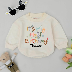 May include: Cream-colored baby onesie with the text "It's my Half Birthday Thomas" in colorful letters. The onesie has long sleeves and a round neckline. Sunglasses and a teddy bear are also in the image.