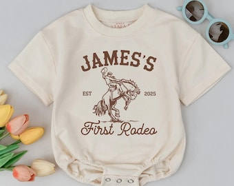 May include: Cream-colored baby bodysuit with short sleeves and snap closures. The front features a brown graphic of a rodeo rider and the text "JAMES'S First Rodeo" with the year "2025". Light blue sunglasses rest on the side.