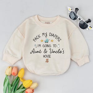 May include: Cream-colored baby bodysuit with the text "Pack My Diapers I'm Going To Aunt & Uncle's House". The design includes a baby carriage, stars, a bottle, and a teddy bear. Sunglasses and flowers are in the background.