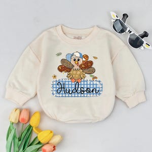 May include: A cream-colored baby bodysuit with a turkey graphic wearing a hat. The turkey has brown and tan feathers and is on a blue gingham name tag that says "Hudson". Sunglasses and flowers are in the background.