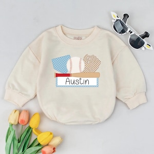 May include: Cream-colored baby sweatshirt with a baseball-themed graphic. The design includes a baseball, bat, and glove, with the name "Austin" displayed on a blue and white label. Sunglasses and flowers are also in the image.