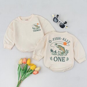 May include: Cream-colored baby outfit set with a long-sleeved sweatshirt and a romper. The sweatshirt has a fishing graphic and the name "Thomas". The romper features a fishing scene with the text "O-FISH-ALLY ONE".