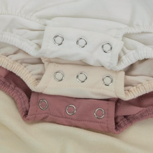 May include: Close-up of three layers of baby clothing in white, cream, and mauve. Each layer has a snap closure with three silver snaps. The fabric appears to be soft and comfortable, with a slight texture.