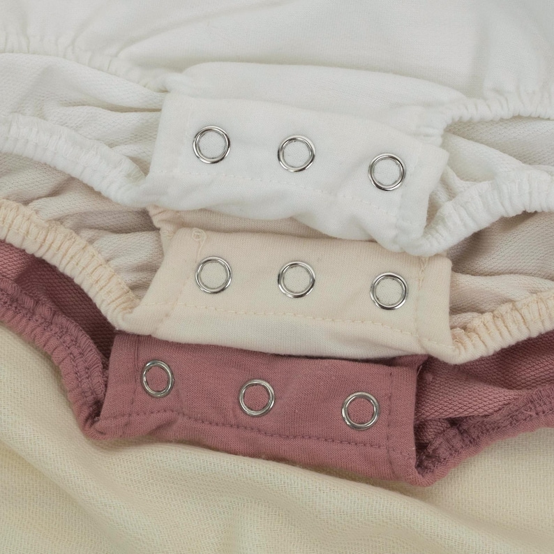 May include: Close-up of three layers of baby clothing in white, cream, and mauve. Each layer has a snap closure with three silver snaps. The fabric appears to be soft and comfortable, with a slight texture.