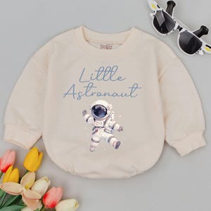 May include: A cream colored sweatshirt with long sleeves features a blue graphic of an astronaut with the text "Little Astronaut" in blue.