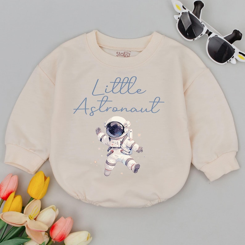 May include: A cream colored sweatshirt with long sleeves features a blue graphic of an astronaut with the text "Little Astronaut" in blue.