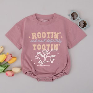 May include: Dusty rose baby romper with short sleeves and a round neckline. The front features the text "ROOTIN' and most definitely TOOTIN'" above a graphic of a cowboy riding a horse. Light blue sunglasses are on the top right.