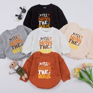 May include: Five long-sleeved baby onesies in different colors with the text "I'll bring the rolls" in orange, yellow, and white. The onesies are on a white background.