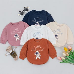 May include: Five long-sleeved shirts with a graphic of an astronaut and the text "Little Astronaut". The shirts are in different colors: pink, white, beige, navy blue, and orange.