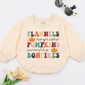 May include: A cream-colored long-sleeved sweatshirt with a colorful graphic print that says "Flannels, Hayrides, Pumpkins, Sweaters, Bonfires". The graphic also includes two orange pumpkins with green stems.