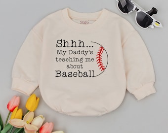 May include: A cream-colored baby onesie with the text "Shhh... My Daddy's teaching me about Baseball" and a baseball graphic. The long-sleeved onesie has a round neckline and is paired with sunglasses and flowers.