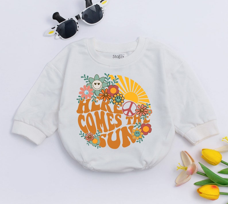 May include: White long-sleeved baby onesie with a graphic print of a sun with flowers and the text "Here Comes the Sun".