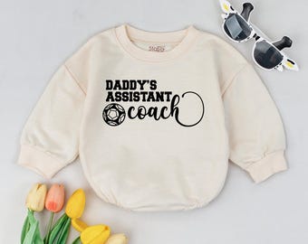 May include: A cream colored long-sleeved sweatshirt with the text "Daddy's Assistant Coach" and a soccer ball graphic.