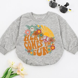 May include: Grey long-sleeved sweatshirt with a retro-style graphic of a sun, flowers, and the text "Here Comes the Sun".