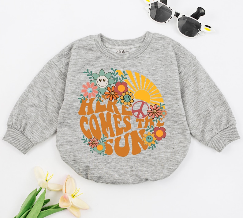 May include: Grey long-sleeved sweatshirt with a retro-style graphic of a sun, flowers, and the text "Here Comes the Sun".