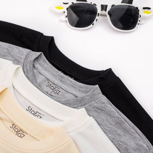 May include: Four crew neck sweatshirts in different colours: beige, white, grey, and black. Each sweatshirt has a small black label with the word "Stafaz" on it.