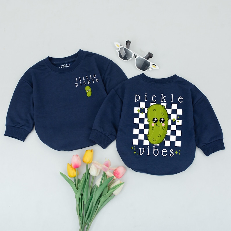 May include: Two navy blue long-sleeved shirts with a pickle graphic. One shirt says "little pickle" and the other says "pickle vibes" with a checkered background.