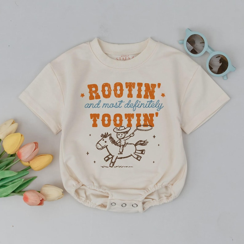 May include: Cream-colored baby onesie with the text "Rootin' and most definitely Tootin'" in orange and blue. The graphic features a cowboy riding a horse. Light blue sunglasses are placed on the side.