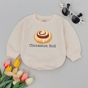 May include: A cream colored long-sleeved sweatshirt with a cinnamon roll graphic and the text "Cinnamon Roll".