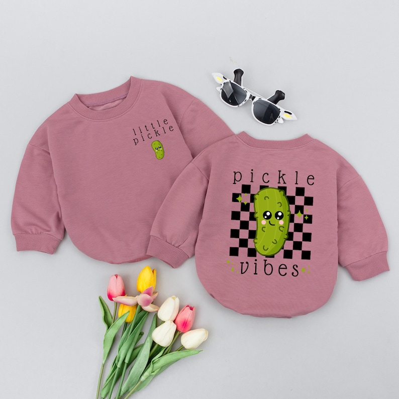 May include: Two pink long-sleeved shirts with a cartoon pickle graphic. One shirt says "little pickle" and the other says "pickle vibes" with a checkered background.