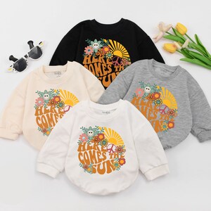 May include: Four long-sleeved shirts with a retro-style graphic print of a sun, flowers, and the text "Here Comes The Sun". The shirts are in different colours: black, beige, white, and grey.