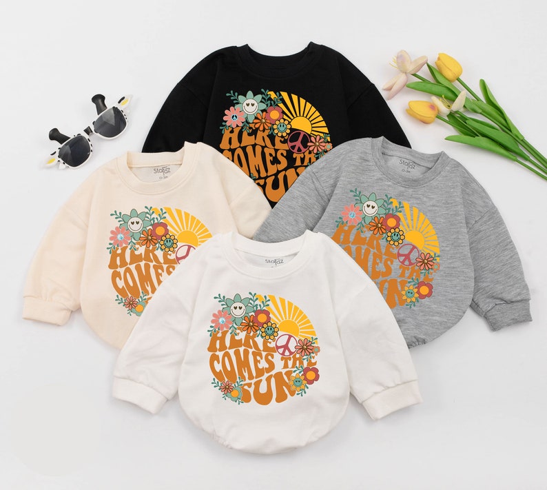 May include: Four long-sleeved shirts with a retro-style graphic print of a sun, flowers, and the text "Here Comes The Sun". The shirts are in different colours: black, beige, white, and grey.