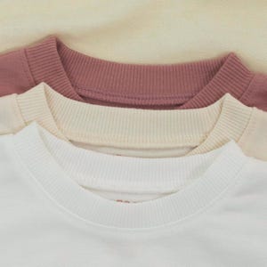 May include: Close-up of three crew neck sweatshirts in white, cream, and mauve. The sweatshirts are stacked, showing the ribbed collar and a portion of the shoulder. The background is a light cream color.