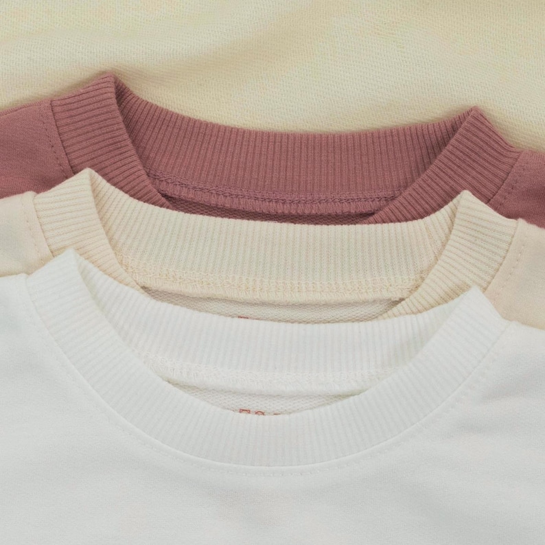 May include: Close-up of three crew neck sweatshirts in white, cream, and mauve. The sweatshirts are stacked, showing the ribbed collar and a portion of the shoulder. The background is a light cream color.