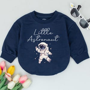May include: A navy blue long-sleeved sweatshirt with a white graphic of an astronaut and the text "Little Astronaut".