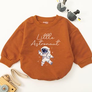 May include: A brown long-sleeved onesie with the text "Little Astronaut" and a white illustration of an astronaut in a spacesuit.