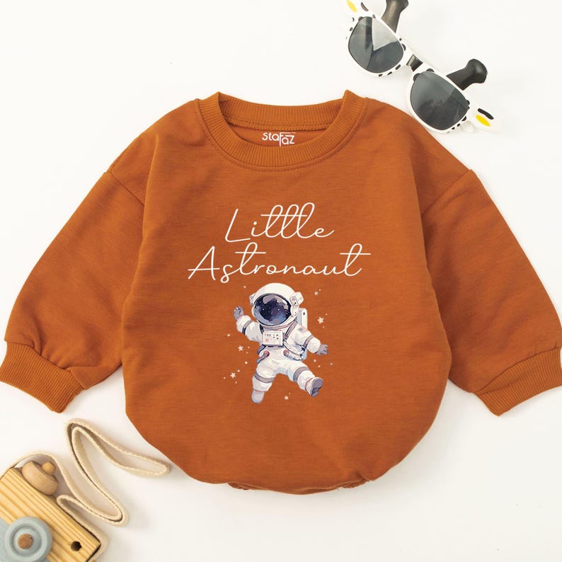 May include: A brown long-sleeved onesie with the text "Little Astronaut" and a white illustration of an astronaut in a spacesuit.