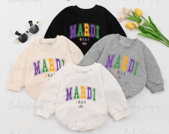 Könnte beinhalten: Vier langärmelige Shirts mit dem Text "Mardi Gras" in grünen, gelben und violetten Buchstaben. Die Shirts sind in Schwarz, Beige, Weiß und Grau.